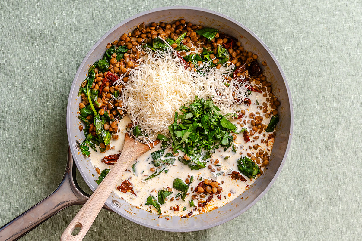 A skillet containing lentils, spinach, and sun-dried tomatoes in a creamy sauce, topped with shredded cheese and fresh herbs, with a wooden spoon resting in the pan.