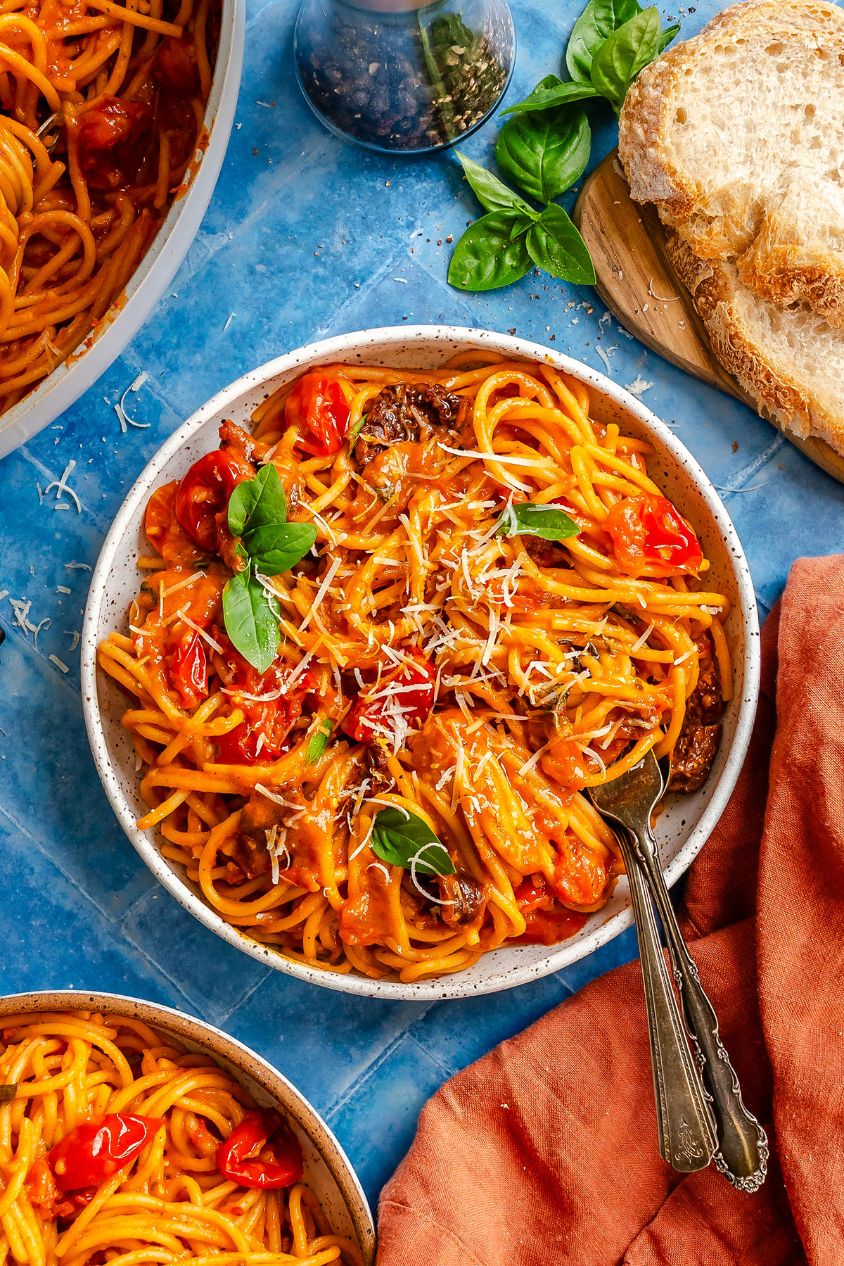 A bowl of tomato basil pasta topped with parmesan cheese, served with crusty bread on a blue surface.