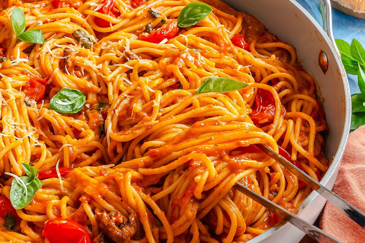 Spaghetti with tomato sauce, cherry tomatoes, and basil leaves in a pan, lifted by metal tongs.