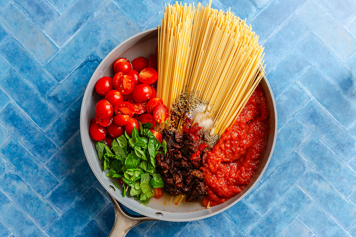 A skillet containing uncooked spaghetti, cherry tomatoes, fresh basil, sun-dried tomatoes, tomato sauce, and seasonings.