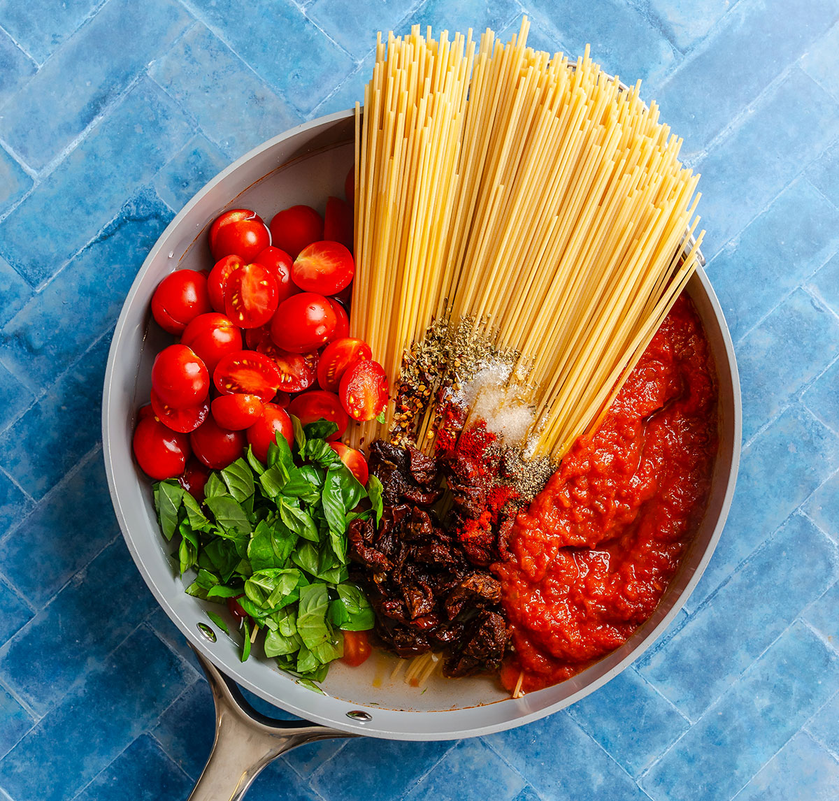 A pan filled with dry spaghetti, cherry tomatoes, tomato sauce, chopped basil, sun-dried tomatoes, and seasonings.