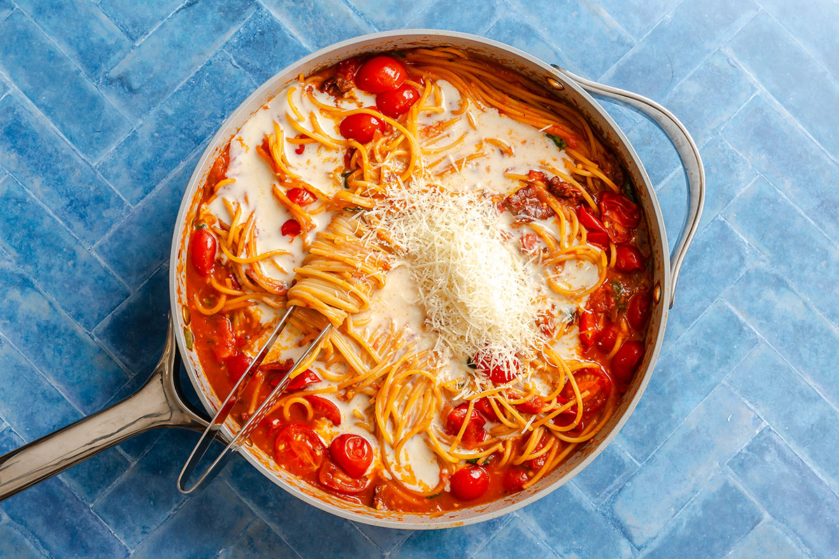 Spaghetti with cherry tomatoes in a creamy sauce, topped with shredded cheese in a metal skillet.