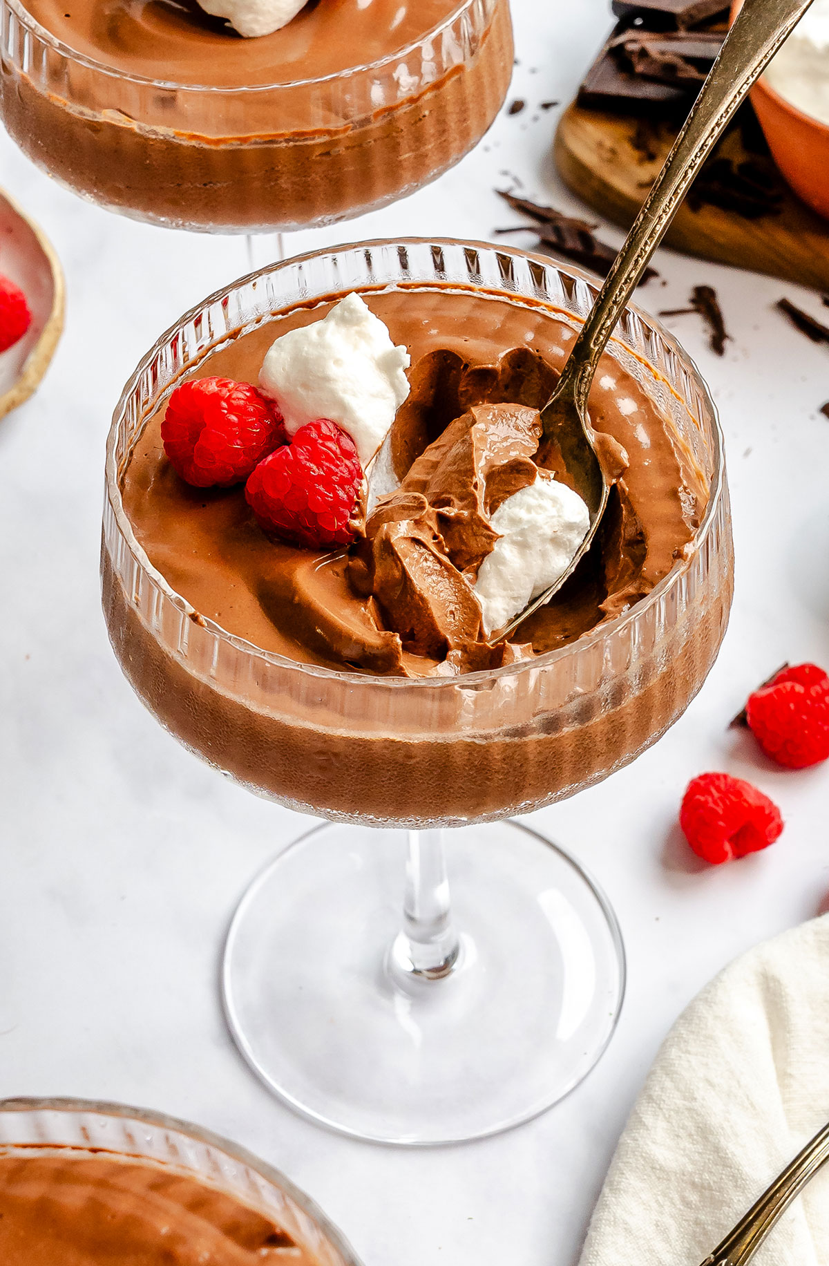 Two glass coupes filled with chocolate mousse topped with whipped cream and fresh raspberries, with a spoon resting in one.
