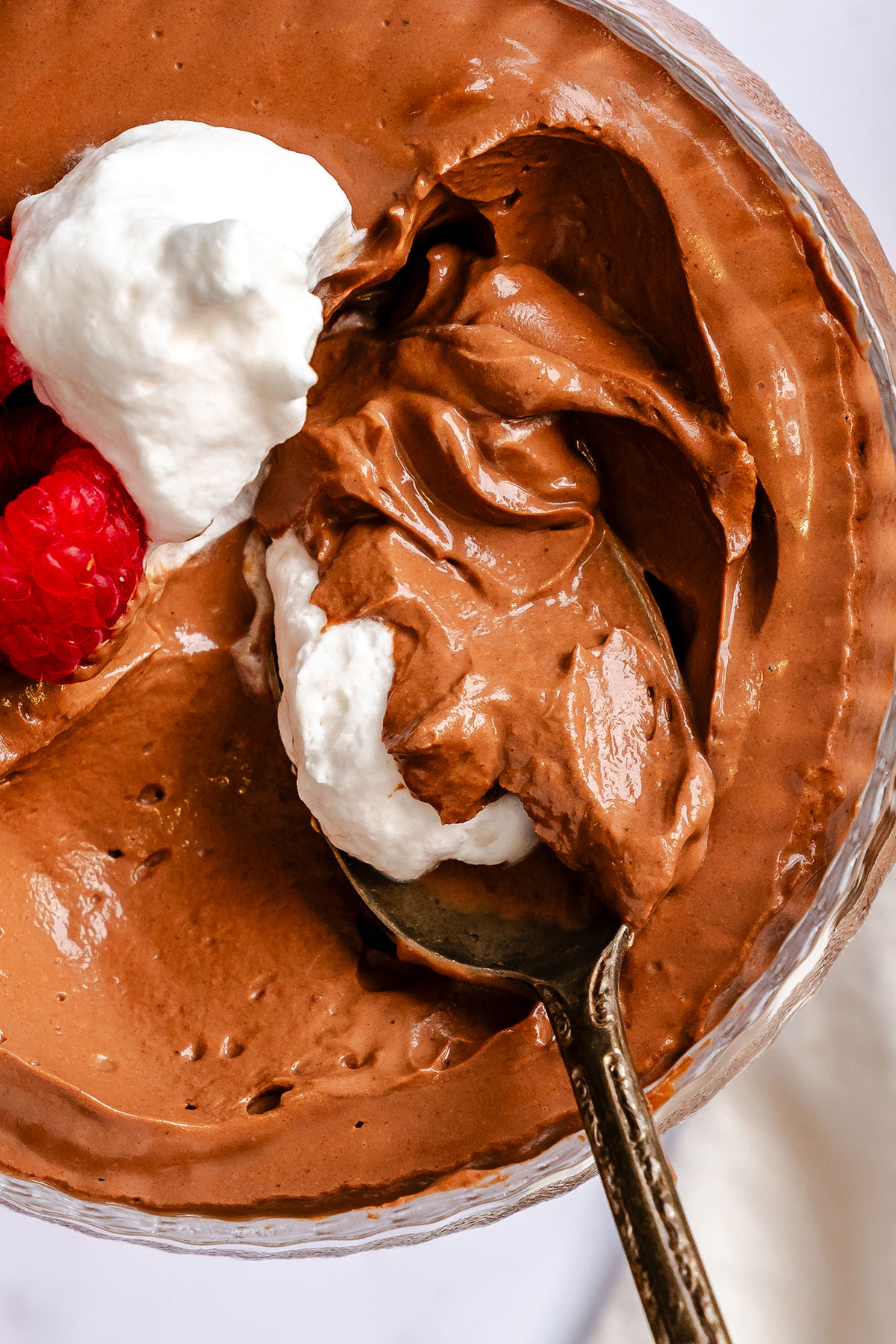 Chocolate mousse in a glass bowl, topped with whipped cream and raspberries, with a spoonful being lifted.