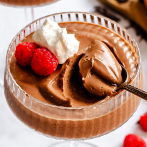 A glass of vegan chocolate mousse topped with whipped cream and two raspberries, with a spoonful being taken out.