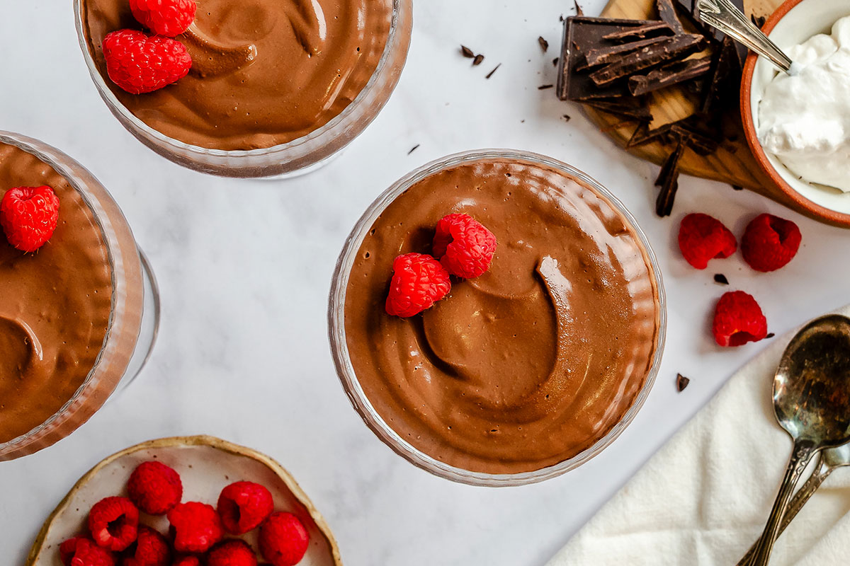 Three glass cups of chocolate mousse topped with fresh raspberries, accompanied by a small bowl of whipped cream, chocolate shavings, and loose raspberries.