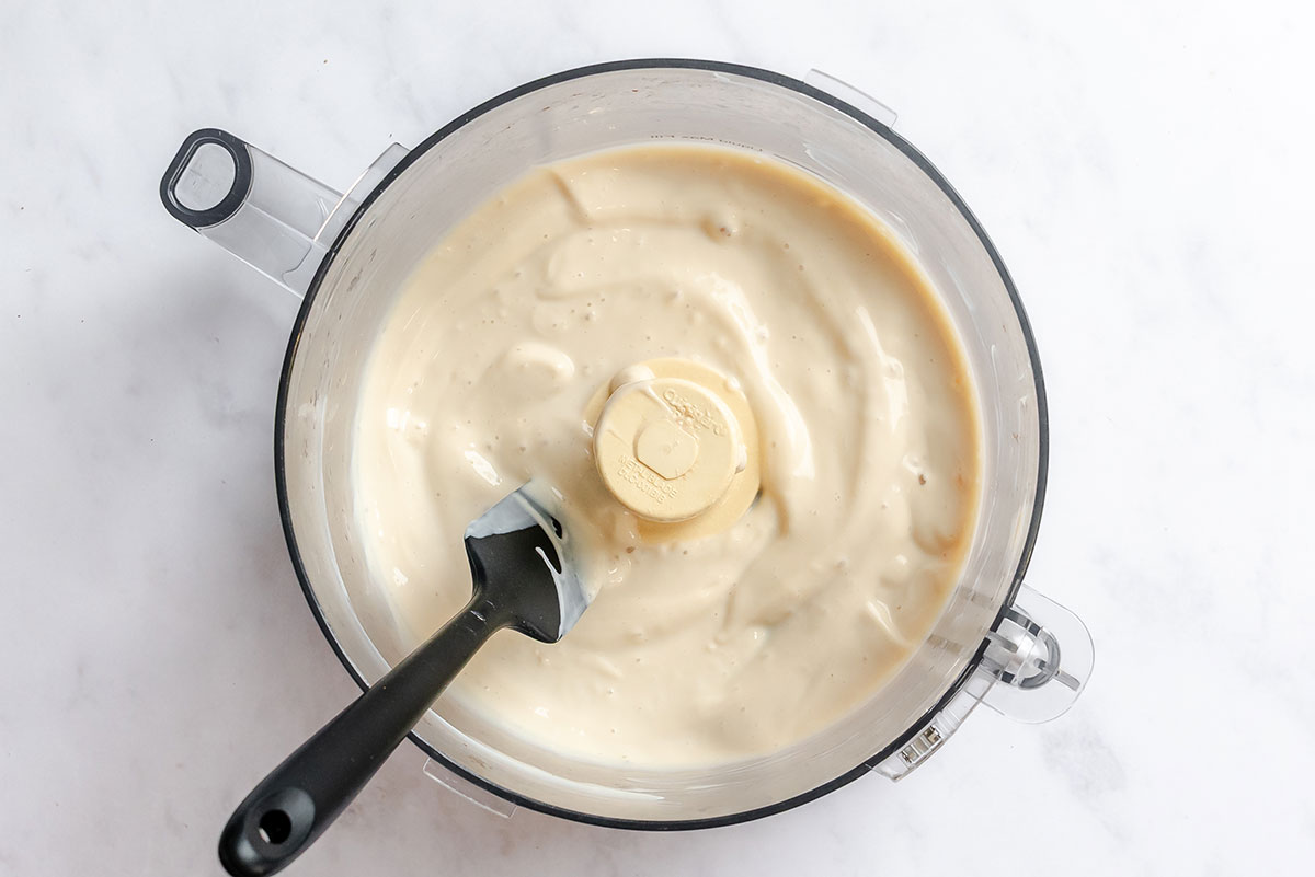 A food processor bowl containing creamy blended sauce and a black spatula.