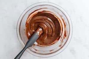 A glass bowl filled with melted chocolate being stirred by a silicone spatula.