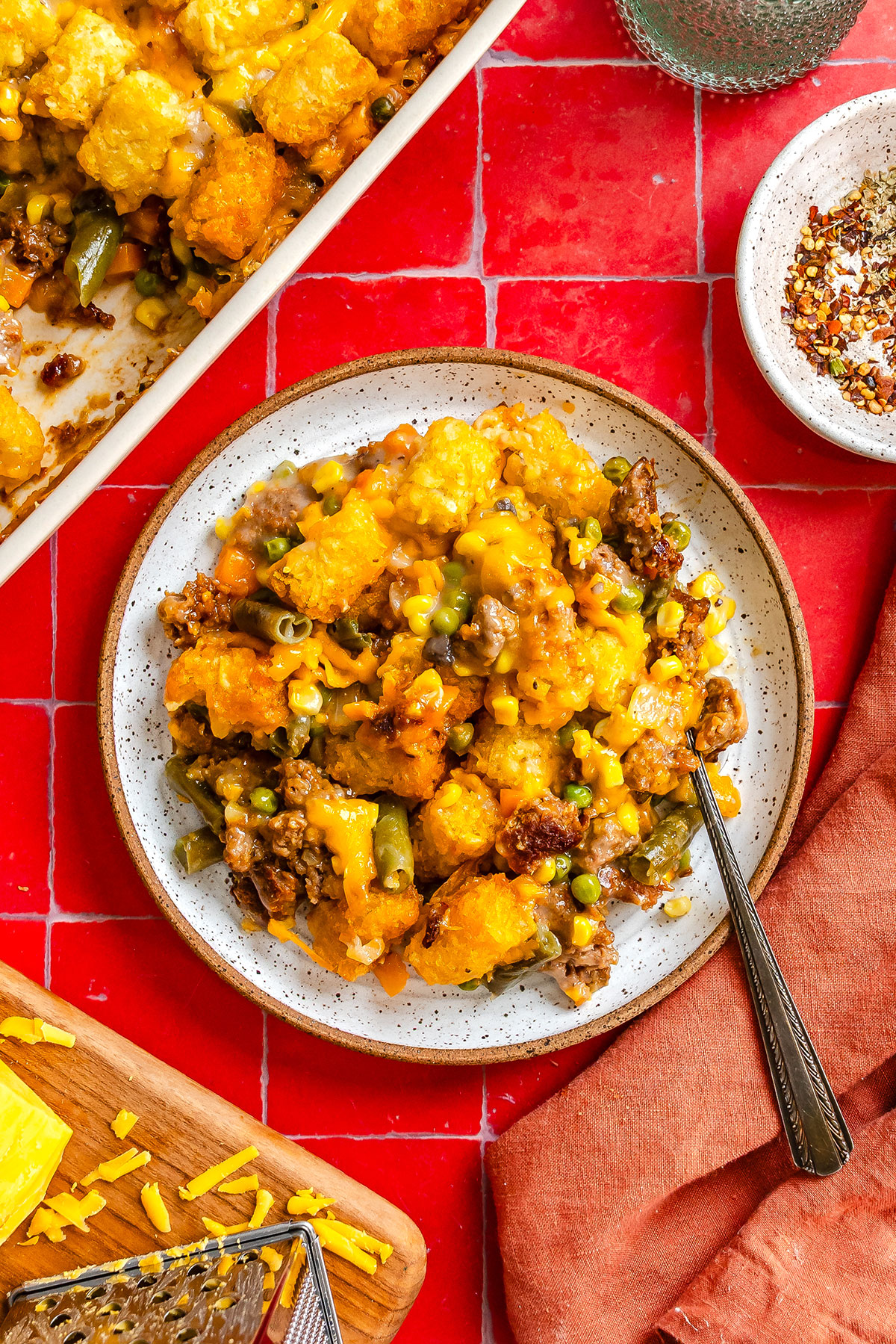 A plate of tater tot casserole with ground meat, corn, and green beans on a red tiled surface.