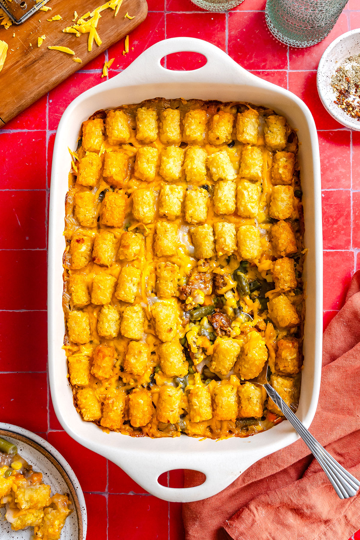 A tater tot casserole with melted cheese in a white baking dish, with a serving spoon resting inside.