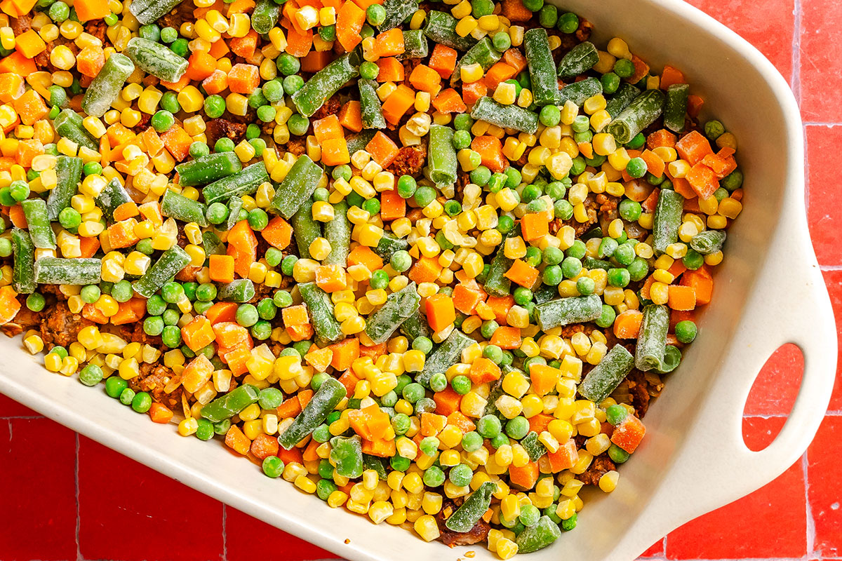 A casserole dish filled with a mixture of ground meat, corn, peas, carrots, and cut green beans.
