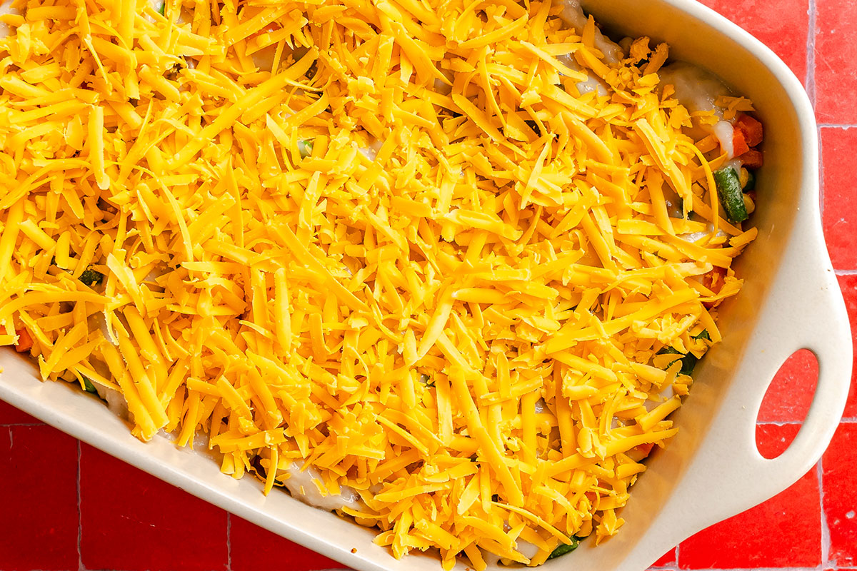 A rectangular baking dish filled with a mixture of vegetables in a creamy sauce, topped with a thick layer of shredded cheddar cheese.